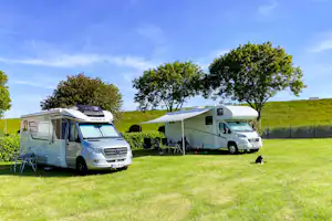 Zwei Wohnmobile auf einem grünen Campingplatz, mit Vorzelten und Stühlen am Stellplatz, im Hintergrund ein grüner Hügel.
