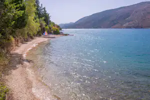 Segelboot am Ufer in einer Campingbucht, Kiesstrand, Wasser mit Wellen, dahinter bewaldete Hänge und Bäume.