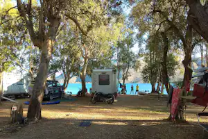 Wohnwagen und Auto auf dem Campingplatz unter Bäumen, mit Blick auf den See und einen Stellplatz am Ufer.