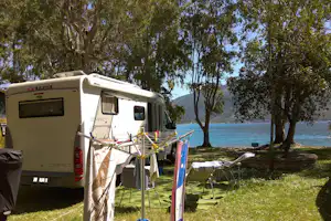 Wohnmobil am Seeufer unter Bäumen, Markise und Campingstühle am Stellplatz, Blick auf das blaue Wasser und Berge.