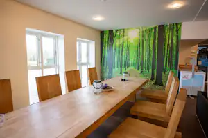 Wohnraum mit Esstisch, Sitzbank und Wandbild mit Waldmotiv, dazu Fenster und Blick ins Freie.