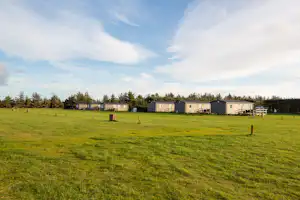 Weite Campingwiese mit Mobilheimen im Hintergrund, niedrigen Bäumen und einzelnen Pfosten im grünen Gras.