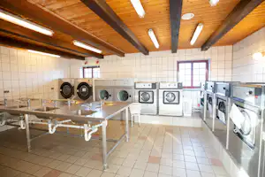 Waschraum im Campingbereich mit Waschbecken, Wäschewagen und mehreren Waschmaschinen an der Wand.