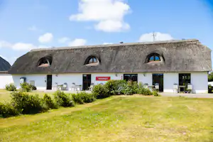Reetgedecktes Ferienhaus mit drei Dachgauben, weißen Türen und Fenstern, davor Rasen und Hecken am Campingplatz.
