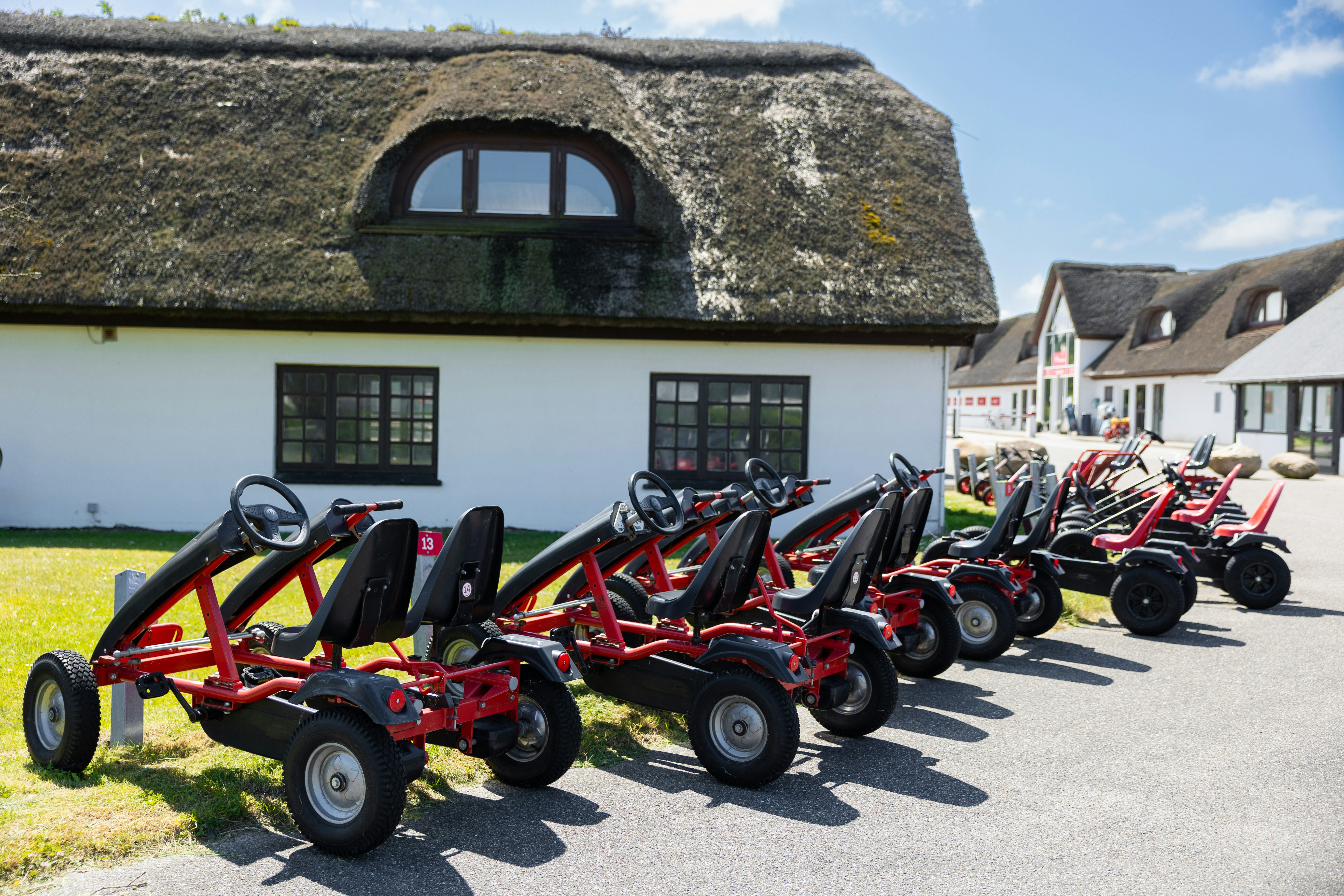 Reihe roter und schwarzer Go-Karts vor einem Reetdach-Haus mit grünen Fenstern und Rasenstreifen am Weg.