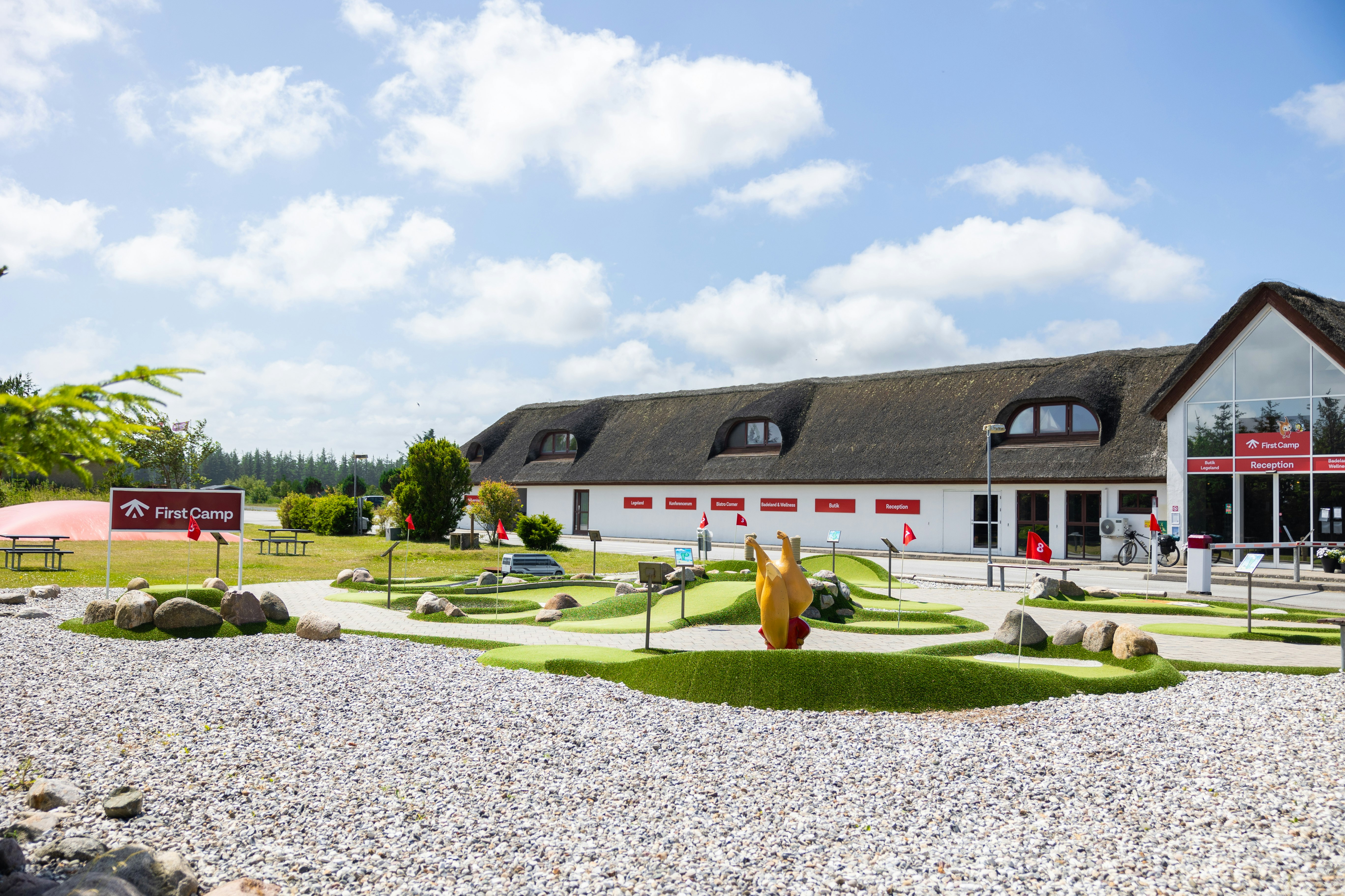 Campinganlage mit gepflegtem Rasen, Kiesweg und einem Gebäude mit mehreren Fenstern und roten Akzenten im Hintergrund.