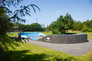 Trampolin auf einem Betonboden in einem Campingplatzbereich, umgeben von Rasen, Bäumen und einem Zaun.