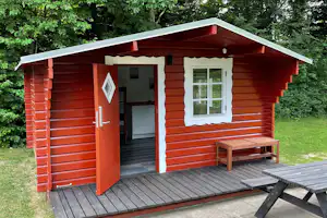 Rote Holzhütte mit Eingangstür, Fenster und Holzbank auf einer Terrasse am Campingplatz, umgeben von grünen Bäumen.