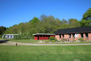 Rasenfläche vor Campinggebäuden mit Ziegelwand, roten Akzenten und Baumreihe im Hintergrund.