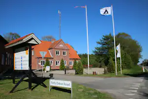 Campingplatzeingang mit Schild „Rally Dakar Road“, Flaggenmasten und Gebäuden mit roten Ziegeldächern am Straßenrand.