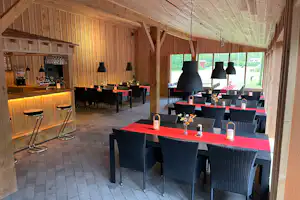 Holz-Essbereich mit großen Tischen, schwarzen Stühlen und Beleuchtung, offene Fensterfront und Fliesenboden im Campinghaus.
