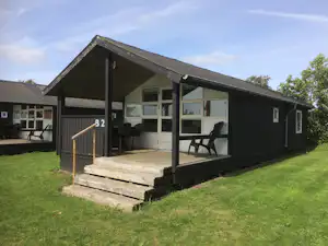Campinghaus mit Terrasse, Sitzgelegenheiten und großen Fenstern, Nummer B2 am Eingang, umgeben von Rasen und Bäumen.