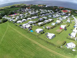 Luftaufnahme eines Campingplatzes mit vielen Wohnwagen und Stellplätzen auf grünen Wiesen, am See und nahe einem Dorf.