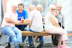 Gruppe von Freunden sitzt auf einer Camping-Terrasse, hält Getränke und lacht vor einem Zelt mit bunten Vorhängen.
