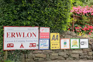Schilder für Caravan- und Camping-Touring Park mit „Welcome To Wales“ vor grüner Hecke und Blumen am Steinmauer.