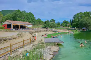 Campingplatz am grünen Fluss mit Terrasse, Steg und Liegewiese; im Wasser sind mehrere Personen zu sehen.