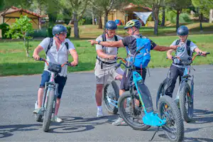 Familie fährt mit Fahrrädern auf dem Campingplatzweg, mehrere Kinder und Erwachsene mit Helmen, Bäume und Rasen im Hintergrund.