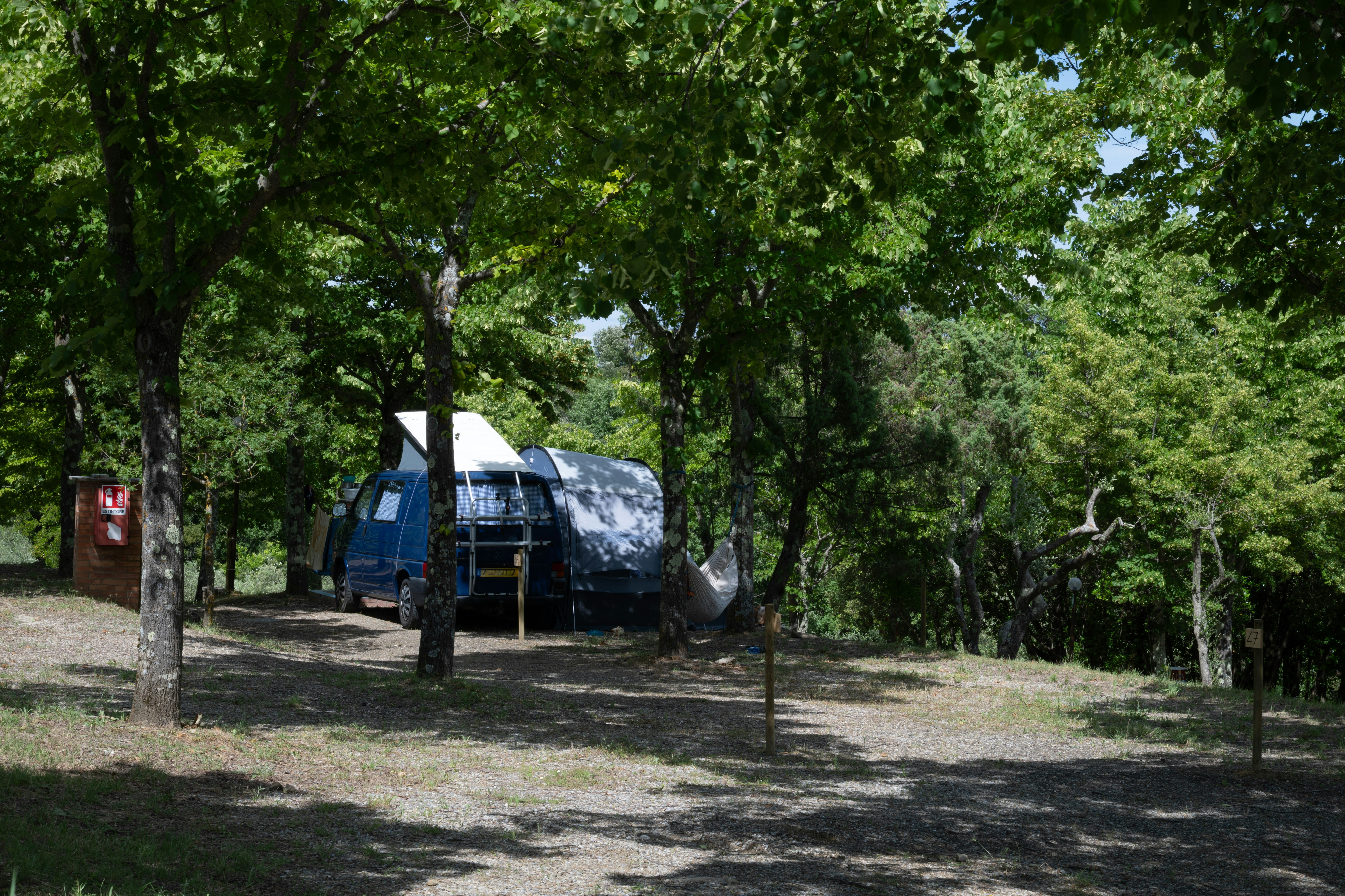 Stellplatz auf dem Campingplatz mit Zelt, Auto und Vorzelt zwischen Bäumen, auf Kiesboden.