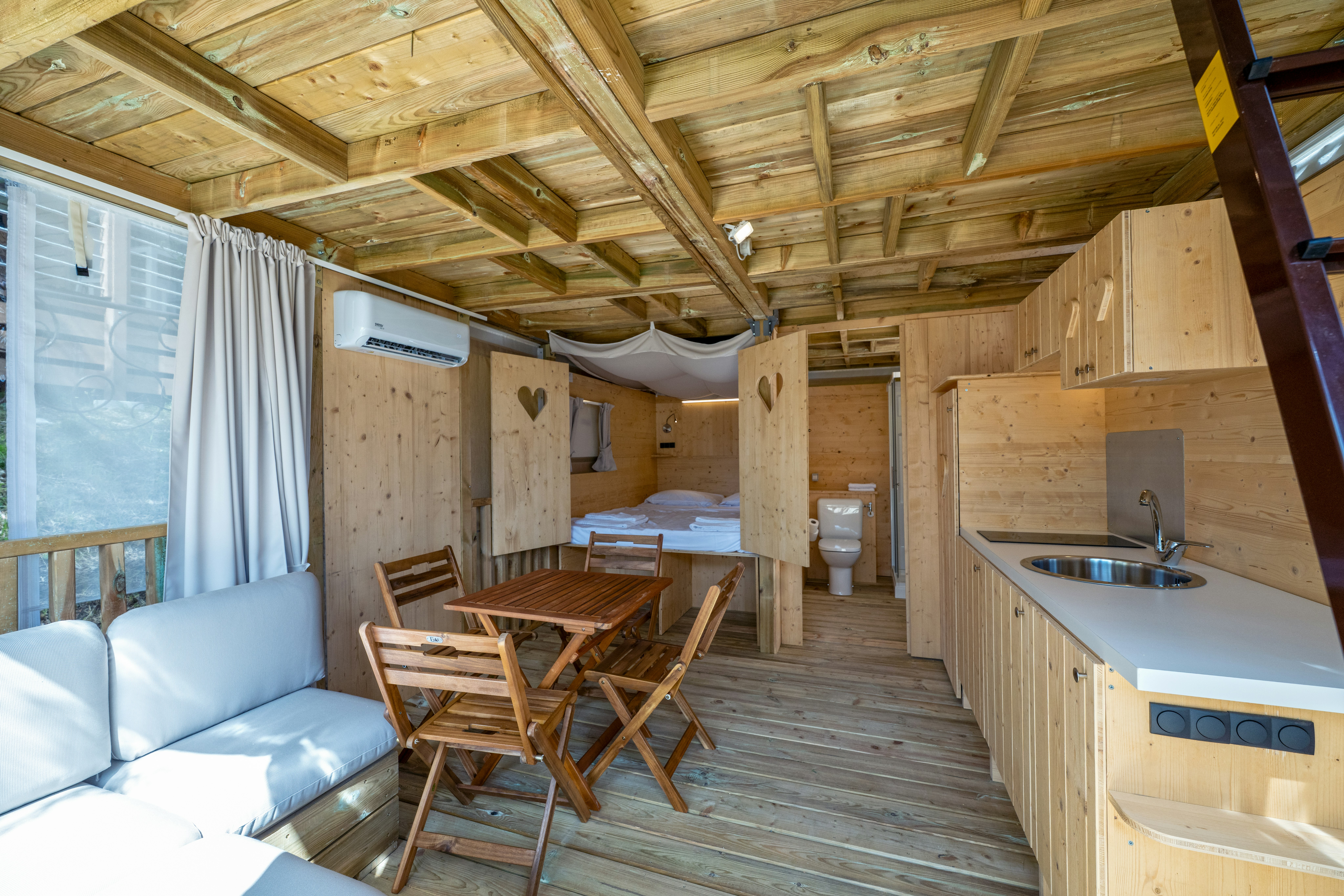 Innenansicht eines Wohnwagens mit Küchenzeile, Esstisch und Stühlen sowie Sofa unter Holzdecke und Vorhang am Fenster.