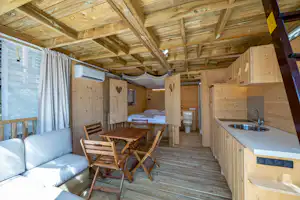 Innenansicht eines Wohnwagens mit Küchenzeile, Esstisch und Stühlen sowie Sofa unter Holzdecke und Vorhang am Fenster.