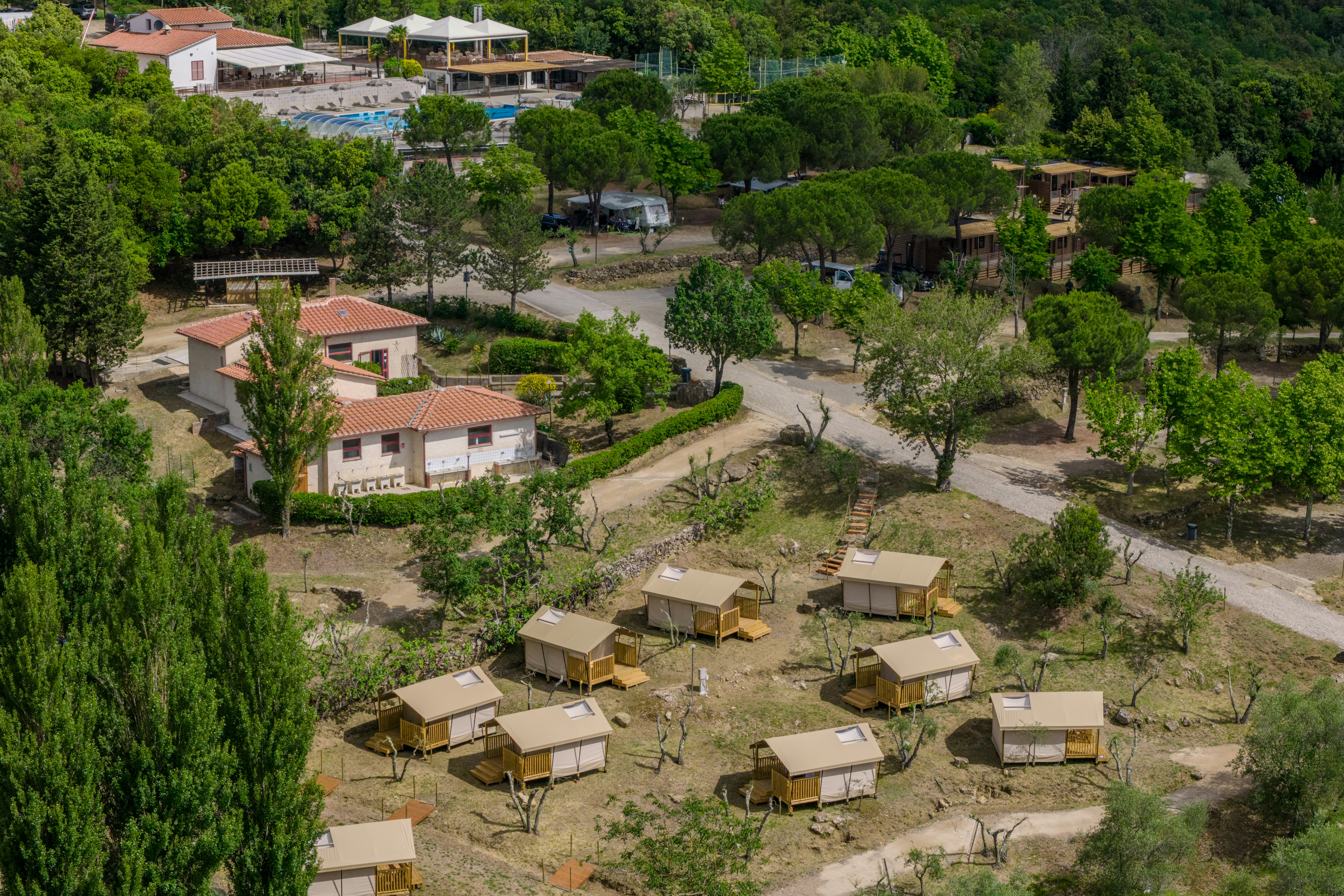 Luftaufnahme eines Campingplatzes mit mehreren Stellplätzen, kleinen Unterkünften, Wegen und Bäumen in hügeliger Landschaft.