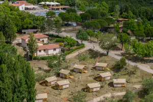 Luftaufnahme eines Campingplatzes mit mehreren Stellplätzen, kleinen Unterkünften, Wegen und Bäumen in hügeliger Landschaft.
