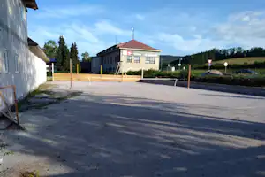 Terrasse am Campingplatz mit Holzgeländer, Betonboden und Blick auf ein Gebäude mit rotem Dach und Bäumen im Hintergrund.