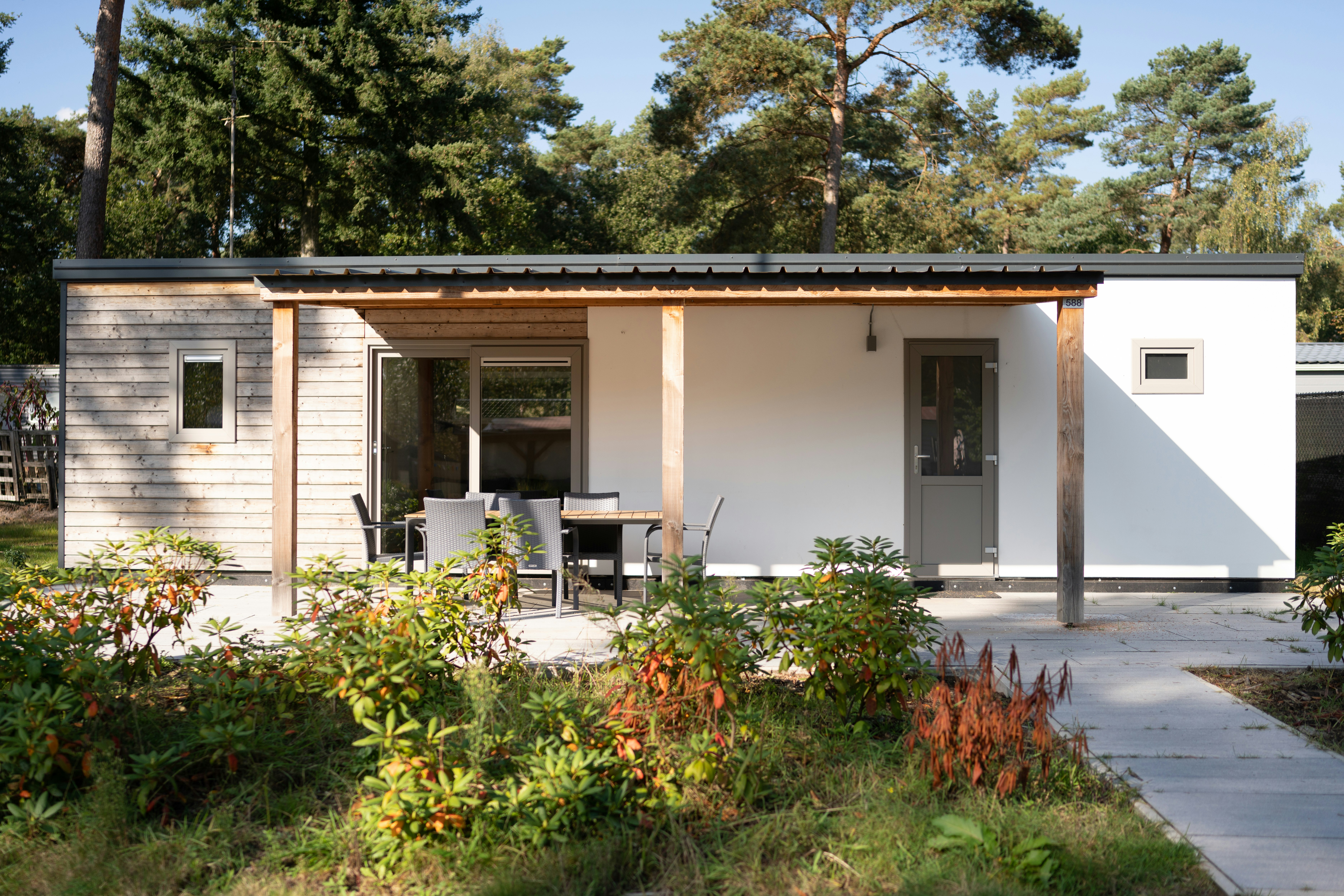 Modernes Ferienhaus mit Terrasse und Sitzbank, umgeben von Büschen und Bäumen, mit gepflastertem Weg am Campingplatz.