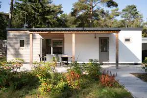 Modernes Ferienhaus mit Terrasse und Sitzbank, umgeben von Büschen und Bäumen, mit gepflastertem Weg am Campingplatz.