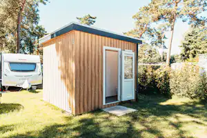 Holz-Mobilheim mit geöffnetem Eingangstürbereich und Vorplatz, daneben ein Wohnwagen auf einem Campingplatz im Grünen.