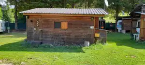 Holzhütte auf einem Campingplatz mit Rasen, Holzfassade, kleinem Fenster und Eingangstür, umgeben von Bäumen.