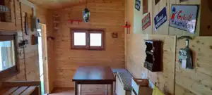 Holzblockhütte innen: Tisch, Bank und Fenster, daneben Küchenzeile mit Wasserhähnen und Schildern an der Wand.