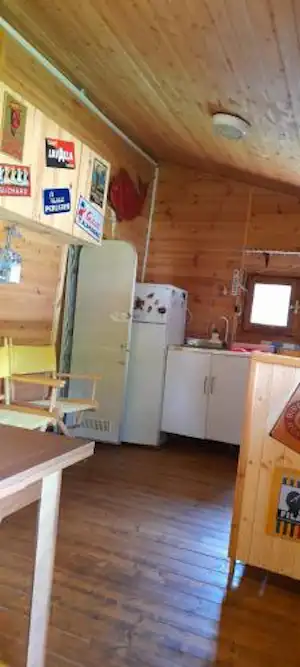 Innenansicht eines kleinen Campinghauses mit Holzdecke, Küchenzeile, Kühlschrank und Fenster, daneben ein Tisch und Stühle.