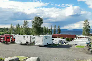 Campingplatz mit Wohnwagen und Wohnmobilen auf Stellplätzen, Bäume und ein See im Hintergrund.