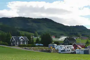 Wohnwagen und Campingfahrzeuge am Rand eines grünen Feldes, dahinter Häuser im Tal und bewaldete Hügel.