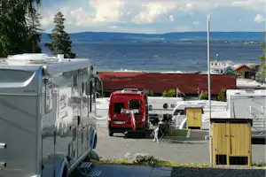 Wohnmobil-Stellplatz mit mehreren Fahrzeugen, Vorzelt und Tisch, Blick auf den See und die Uferpromenade.