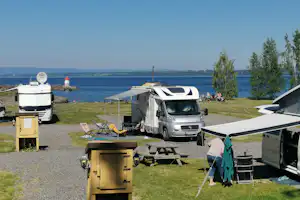 Wohnmobil und Zelte auf einem Campingplatz am See, mit Picknicktisch, Stühlen und Blick auf das Wasser.