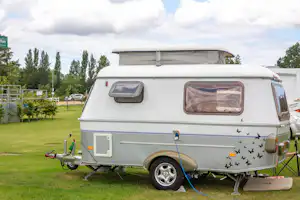 Wohnwagen auf einer Campingwiese mit Vorzelt, Anhänger und blauem Schlauch, im Hintergrund Bäume und ein Parkplatz.