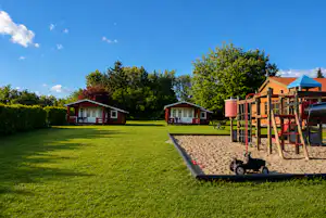 Spielplatz mit Holzgerüst und Sandfläche auf grünem Rasen, im Hintergrund mehrere Ferienhäuser und Bäume.