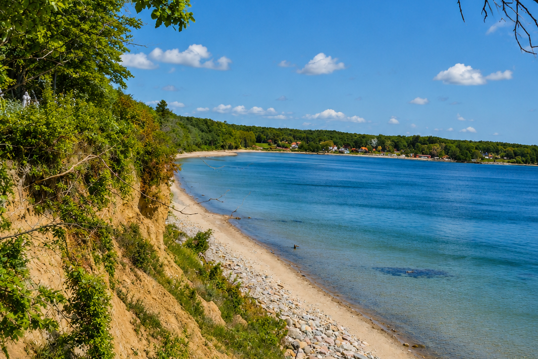 Blick auf den See mit sandigem Ufer, Kiesstrand und Bäumen am Hang unter blauem Himmel mit Wolken.