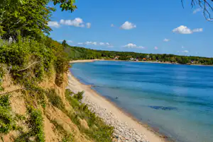 Blick auf den See mit sandigem Ufer, Kiesstrand und Bäumen am Hang unter blauem Himmel mit Wolken.