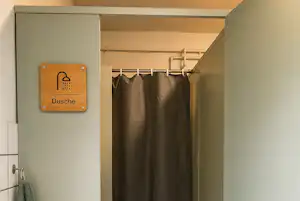 Duschraum mit Duschvorhang, Handtuch an der Wand und Fliesenboden, daneben eine Tür und Sanitärarmaturen.