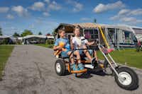 Familiecamping De Molenhoek -  Kinder auf einem Kettcar mit dahinterliegenden Stellplätzen 