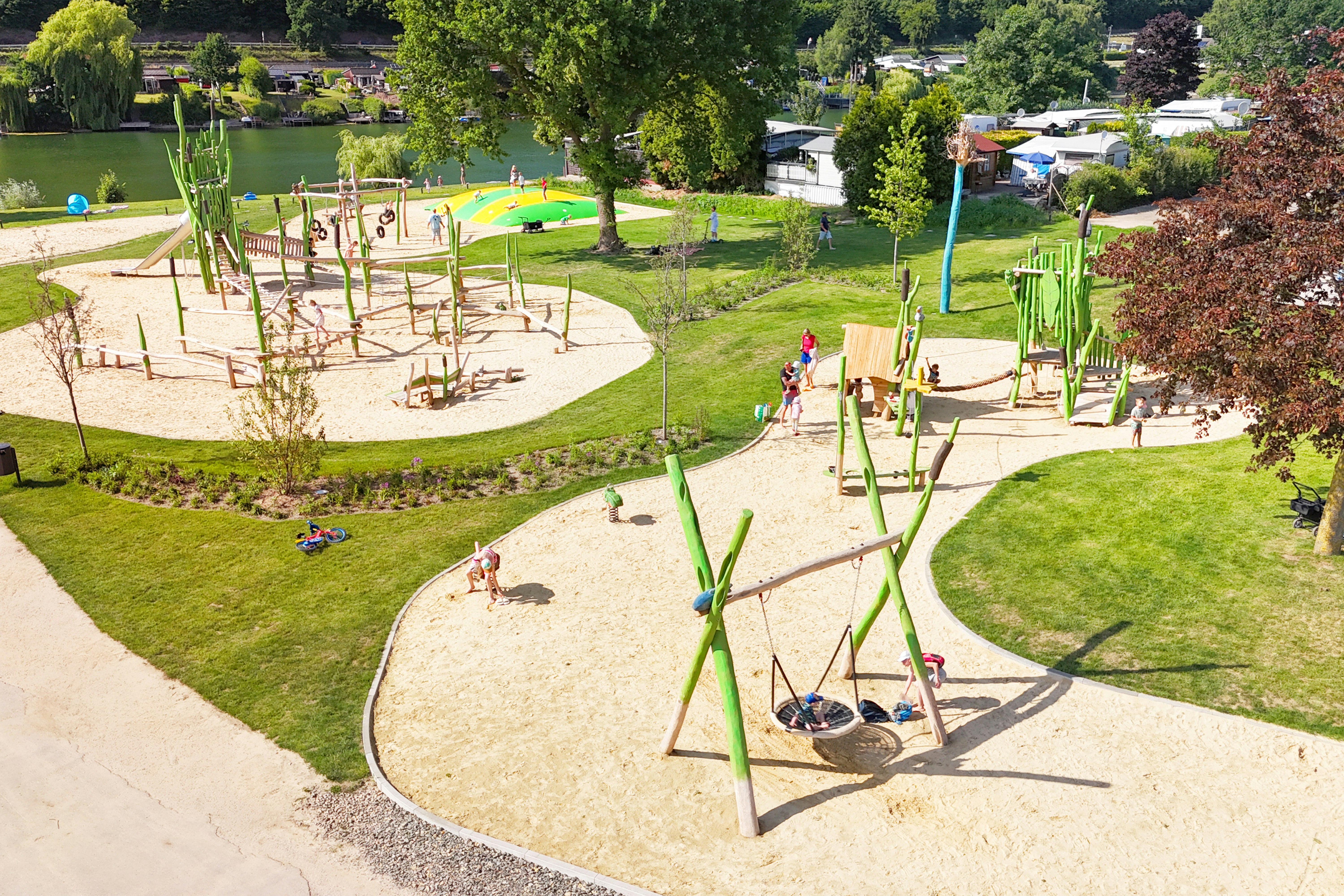 Spielplatz im Campinggarten mit Klettergerüst, Sandfläche und Wegen, im Hintergrund Wohnwagen und Bäume.