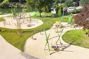 Spielplatz im Campinggarten mit Klettergerüst, Sandfläche und Wegen, im Hintergrund Wohnwagen und Bäume.