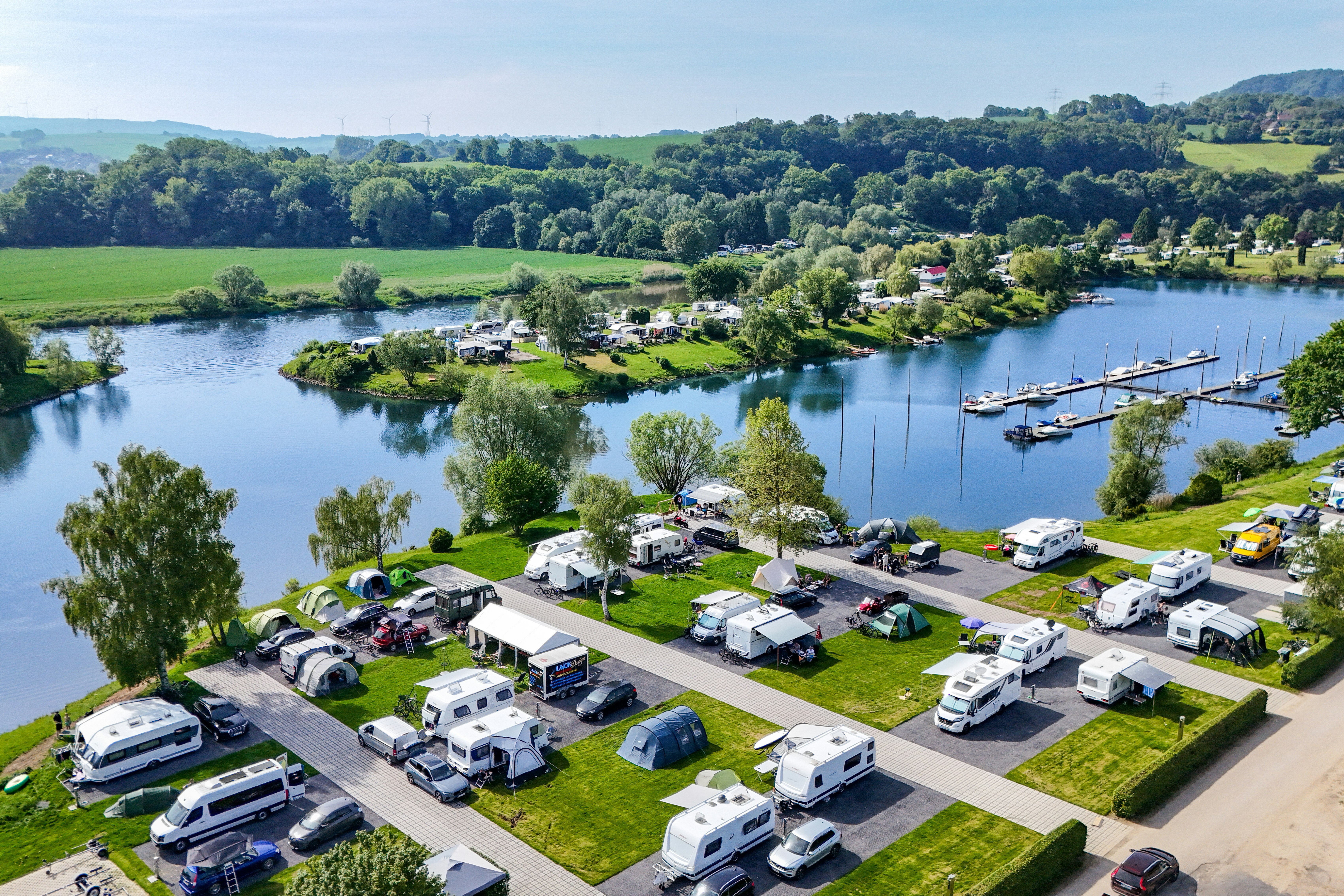 Campingplatz am See mit Stellplätzen, Wohnmobilen und Wohnwagen, Booten im Wasser und Blick auf die grüne Landschaft.