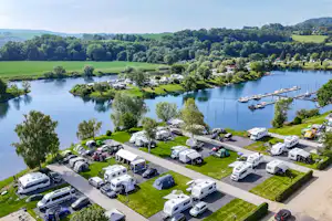 Campingplatz am See mit Stellplätzen, Wohnmobilen und Wohnwagen, Booten im Wasser und Blick auf die grüne Landschaft.