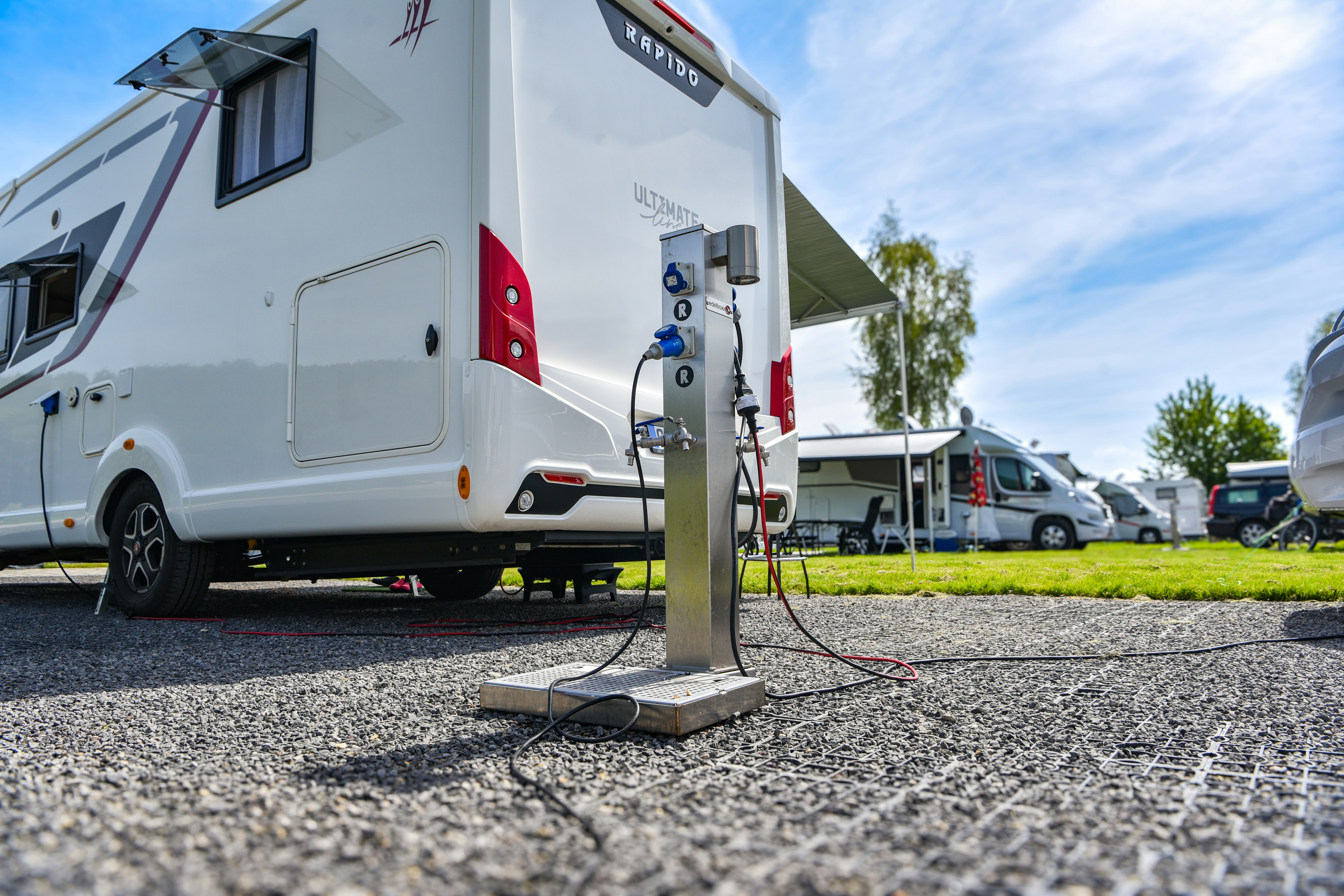 Wohnmobil am Stellplatz mit Stromsäule, Kiesboden und Wohnwagen im Hintergrund, Zelte und Bäume neben dem Campingplatz.