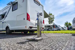 Wohnmobil am Stellplatz mit Stromsäule, Kiesboden und Wohnwagen im Hintergrund, Zelte und Bäume neben dem Campingplatz.