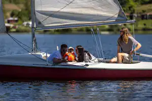 Segelboot auf dem Wasser: Familie sitzt im Boot, Kinder schauen nach vorn, im Hintergrund Ufer und Bäume.